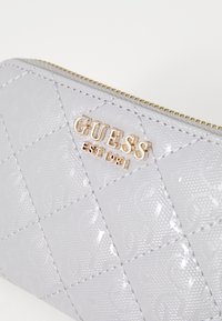 Grijze, gewatteerde synthetische portemonnee met een glanzende afwerking, een gouden "GUESS"-logo en een reliëfdetail van het oprichtingsjaar 1981. Ritsluiting.