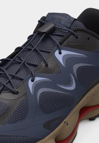 Granatowo-czarny but sportowy z wzorzystą siateczkową cholewką, elastycznymi sznurowadłami zabezpieczonymi stoperem oraz beżową podeszwą z czerwonym akcentem.