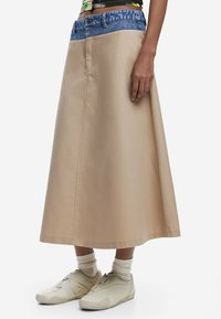 Jupe midi beige avec une taille en denim, présentant une texture lisse et des lignes simples. Associée à des chaussures beige clair et des chaussettes crème.