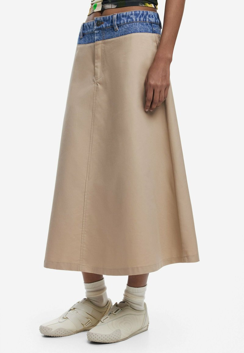 Jupe midi beige avec une taille en denim, présentant une texture lisse et des lignes simples. Associée à des chaussures beige clair et des chaussettes crème.