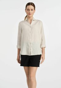 Jasno beżowa lniana koszula zapinana na guziki z długimi rękawami, kołnierzykiem i prostym dołem, w zestawieniu z czarnymi szortami. Prosty design i tekstura.