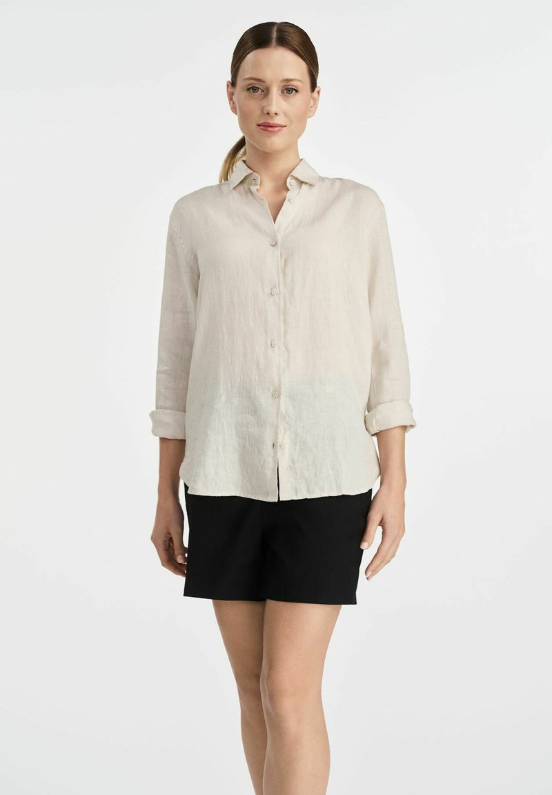 Jasno beżowa lniana koszula zapinana na guziki z długimi rękawami, kołnierzykiem i prostym dołem, w zestawieniu z czarnymi szortami. Prosty design i tekstura.