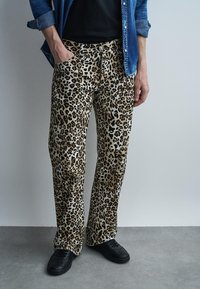 Άτομο που φοράει μπεζ και καφέ παντελόνια με λεοπάρ σχέδιο, μαύρα παπούτσια, μαύρο πουκάμισο και μπλε τζιν πουκάμισο με κουμπιά και γυρισμένα μανίκια.