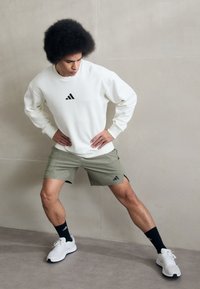 Baltas džemperis ar melnu Adidas logo, olīvzaļas šortus un baltas sporta kurpes ar melniem akcentiem un garām melnām zeķēm.