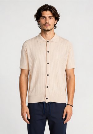 Jonge man met middellang donker haar, gekleed in een beige kortebarms overhemd met knopen en marineblauwe broek, staand tegen een effen achtergrond.