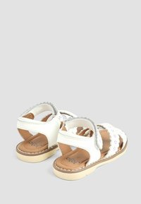 Sandali bianchi in pelle con dettagli scallop, cinturino regolabile e soletta beige. Presentano una suola piatta con una base in gomma chiara.