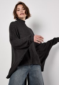 Femme portant un pull noir ample à col roulé avec de larges manches et un jean bleu, posant devant un fond clair et uni.