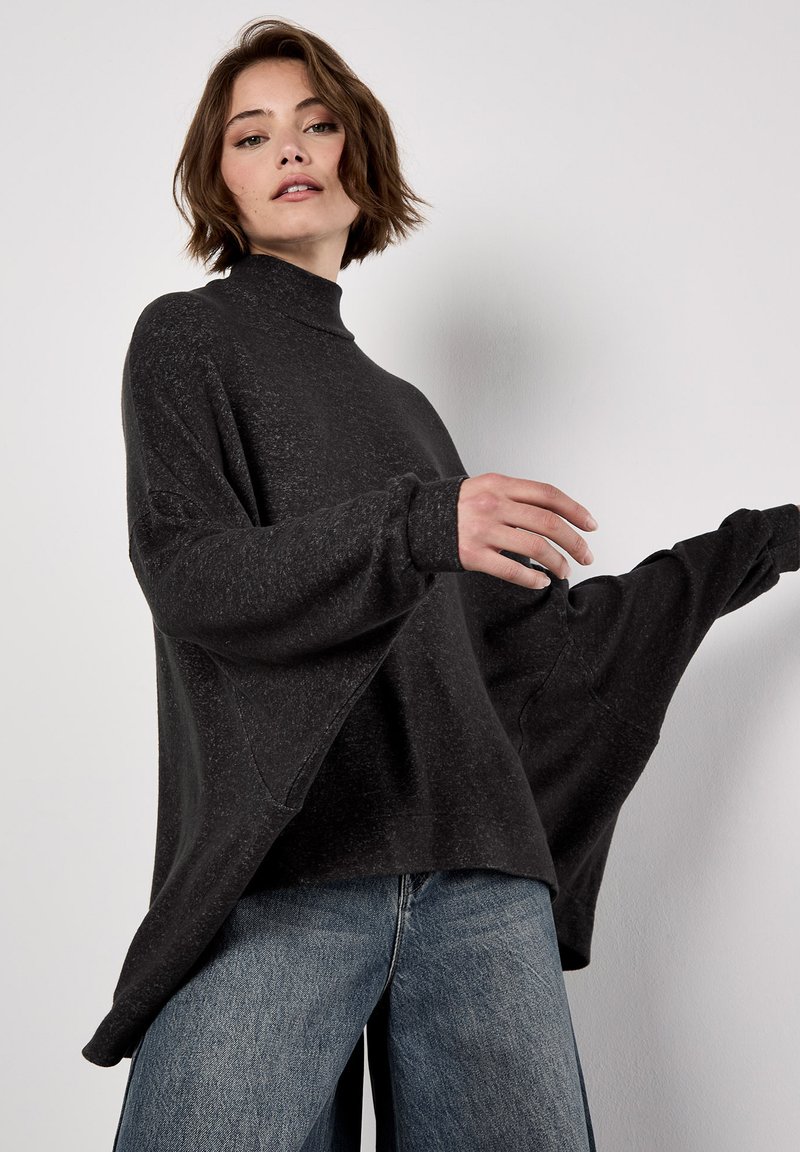 Femme portant un pull noir ample à col roulé avec de larges manches et un jean bleu, posant devant un fond clair et uni.
