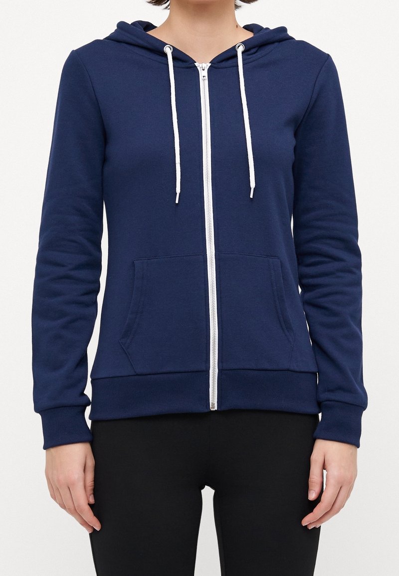 Femme portant un sweat à capuche bleu marine zippé avec cordons blancs et poches avant, assorti à un pantalon noir.