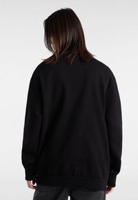 Černá mikina s volným střihem, kulatým výstřihem a dlouhými rukávy. Hladká textura a žebrované manžety. Nošena s tmavě šedými kalhotami.