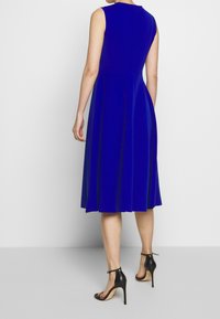 Blå, ärmlös A-linje klänning med en åtsittande överdel, utsvängd kjol och vertikala sömmar. Bärs med svarta, remmade högklackade sandaler.