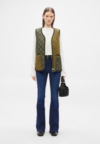 Γιλέκο με γέμιση σε σκούρο πράσινο και ελιά, με φερμουάρ, δύο μπροστινές τσέπες και χρυσό λογότυπο. Συνδυάζεται με καμπάνες μπλε τζιν και μαύρη τσάντα.