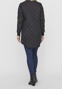 Cappotto lungo trapuntato nero con colletto tondo, polsini a coste e un orlo posteriore più lungo, abbinato a jeans blu aderenti e stivaletti neri.