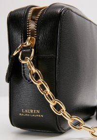 Borsa a tracolla in pelle nera con finitura testurizzata, hardware in oro e tracolla a catena. Presenta una chiusura con zip e un marchio impresso.