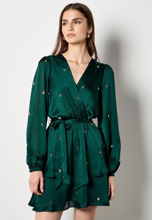 Groene wikkeljurk van gladde stof, met lange mouwen, een strik taille en verspreide gouden paillettenversieringen.