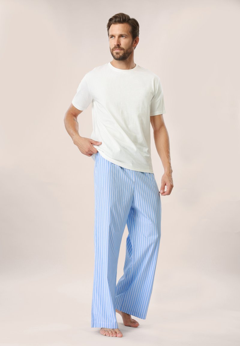 Homme debout pieds nus portant une chemise blanche à manches courtes et un pantalon ample rayé verticalement bleu et blanc sur un fond uni.