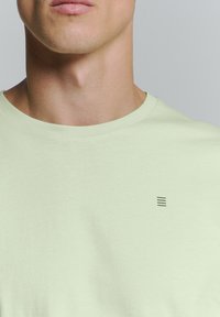Lichtgroene katoenen t-shirt met ronde hals, voorzien van een klein donker grafisch detail aan de linkerkant. Zachte textuur en casual ontwerp.