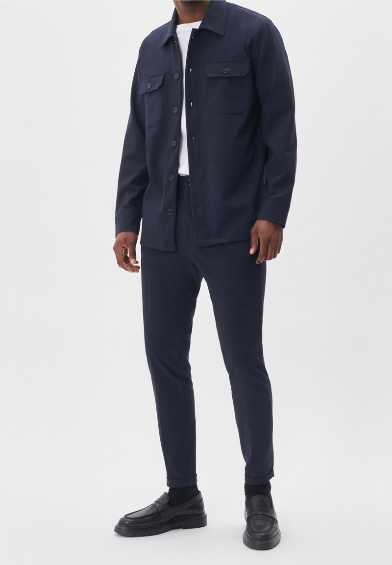 Homme portant une veste bleu marine boutonnée avec poches poitrine, un pantalon slim assorti, des chaussettes noires, des mocassins noirs, et une chemise blanche en dessous.