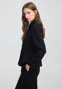 Žena s dlouhými hnědými vlasy, oblečená v přizpůsobeném černém blejzru a odpovídajících kalhotách, stojící v profilu s jednou rukou v kapse na jednobarevném pozadí.