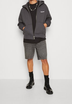Mann trägt dunkelgrauen Kapuzenpullover mit Reißverschluss, schwarzes T-Shirt, graue Jeans-Shorts, schwarze Socken und schwarze Schnürstiefel, steht vor hellem Hintergrund.