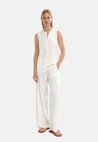 Biała bezrękawnikowa bluzka z zamkiem z przodu, zestawiona z szerokimi białymi spodniami. Uzupełniona czerwonymi klapkami. Gładki materiał i luźny krój.