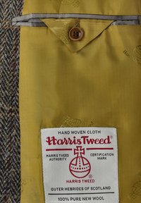 Harris Tweed Etikett auf gelbem Stoff, das einen Knopfverschluss zeigt, mit einem Muster dunklerer Fäden und einem Zertifizierungszeichen für Authentizität.