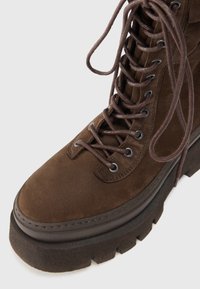 Stivale in suede marrone con lacci, punta arrotondata, superficie testurizzata, occhielli in metallo e suola piatta dal design chunky con motivo a zigzag.