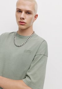 Tricou verde deschis din bumbac, cu mâneci scurte, având un logo brodat subtil. Asortat cu o brățară groasă din argint.
