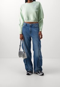 Felpa cropped verde menta, jeans cargo blu a vita alta con tasche e una borsa a tracolla argento. Le sneakers in bianco e nero completano il look.