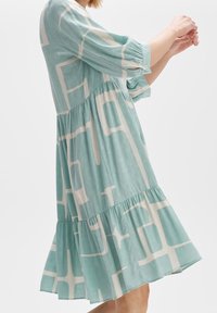 Vaaleansininen mekko, jossa valkoiset geometriset kuviot, pitkät hihat, kerroksellinen helma ja teksturoidun tuntuinen kangas.