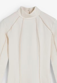 Top de manga larga con cuello tipo tortuga en tejido texturizado blanco roto, que presenta detalles de costura y un cierre de botón en la parte posterior del cuello.