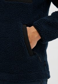Marineblauwe fleece pullover met een gestructureerd oppervlak, zwarte accenten en een zijzak. Heeft een trekkoord bij de zoom voor een aanpasbare pasvorm.