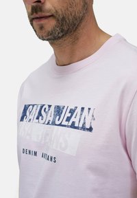 Světle růžová mikina s kulatým výstřihem, která má opotřebovaný grafický potisk s nápisem "SALSA JEAN" v modré barvě a pod ním "DENIM ARTISANS" v černé barvě.
