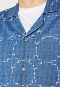 Modrá krátkosleevá košile z denimu s bílým geometrickým vzorem a dřevěným knoflíkem u límce.