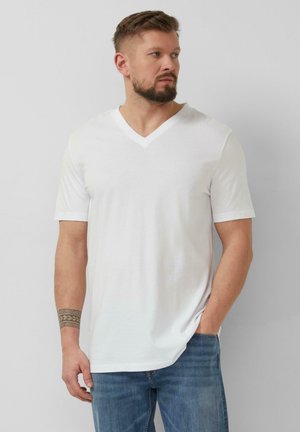 Mężczyzna z brodą, ubrany w biały T-shirt z krótkim rękawem i dekoltem w serek oraz niebieskie jeansy, patrzący na bok, jedną ręką w kieszeni, z tatuażem na przedramieniu.