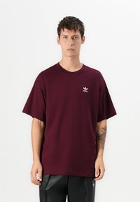 Jauns vīrietis ar īsiem tumšiem matiem, vilkws burgundijas krāsas Adidas t-kreklu un melnas bikses, stāv uz vienkārša balta fona.