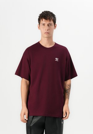 Mlad muškarac s kratkom tamnom kosom koji nosi bordo Adidas majicu i crne hlače, stoji naslonjen na jednostavnu bijelu pozadinu.