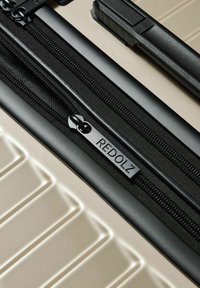 Close-up van een beige harde koffer met een zwarte rits en een trekband, waarin de merknaam "REDOLZ" is gedrukt.