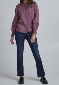 Camicia a maniche lunghe e bottoni, di un lavanda tenue, con colletto e polsini arricciati. Abbinata a jeans a gamba larga blu scuro e scarpe nere lucide.