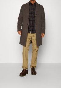 Cappotto in misto lana marrone con colletto a punte, abbinato a una camicia a quadri blu navy e pantaloni beige. Scarpe in pelle marrone completano l'outfit.