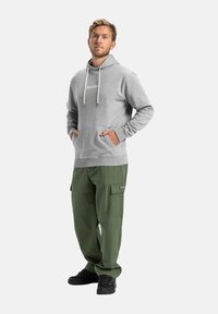 Hoodie gris avec poches avant et cordons blancs, associé à un pantalon cargo vert avec poches latérales et un écusson de logo. Baskets noires.