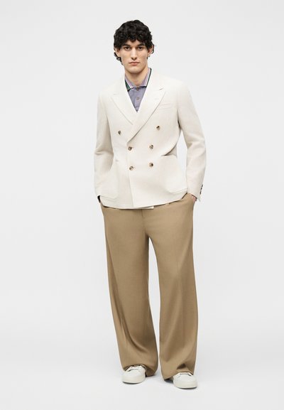 Jeune homme portant un blazer croisé crème, une chemise à col rayée, un pantalon large beige et des baskets blanches, debout les mains dans les poches.