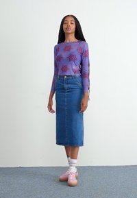 Haut à manches longues violet avec un motif floral, jupe crayon en denim bleu, et baskets roses. Chaussettes blanches visibles, se tenant contre un fond neutre.
