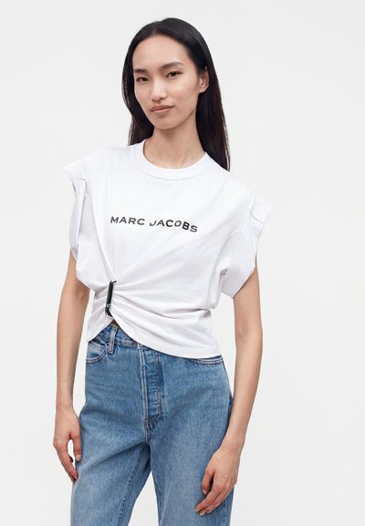 Femme aux longs cheveux foncés portant un t-shirt blanc Marc Jacobs attaché sur un côté et un jean bleu, debout devant un fond uni.