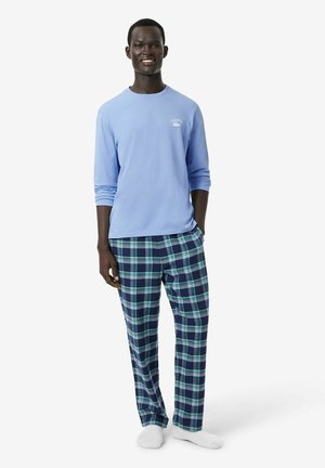 Mann trägt hellblaues Langarmhemd und blau-grüne karierte Pyjamahose, steht und lächelt mit einer Hand in der Tasche vor weißem Hintergrund.
