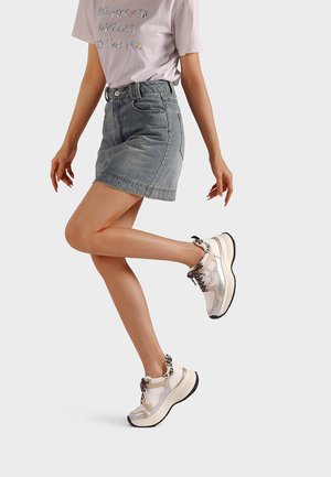 Donna che indossa una gonna di jeans, una T-shirt stampata lilla chiaro e sneakers beige con dettagli animalier, posando con una gamba sollevata.