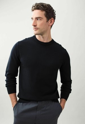 Schwarzer Strickpullover mit Rundhalsausschnitt und gerippten Bündchen, kombiniert mit grauen Hosen. Der Stoff wirkt weich, glatt und leicht.
