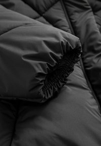 Schwarze Daunenjacke aus einem gequilteten Stoff. Der Ärmelbündchen hat ein elastisches Band und eine weiche Oberfläche, akzentuiert mit glattem schwarzen Material.
