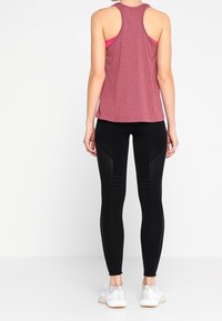 Maroon racerback-top yhdistetty mustiin legginsseihin, joissa on leikattuja kuvioita ja teksturoituja paneeleja. Mallilla on jalassaan valkoiset urheilukengät.