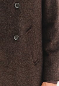 Brązowy wełniany płaszcz z teksturowanej tkaniny, z dwoma guzikami z przodu i kieszonką boczną dla dodatkowego szczegółu.